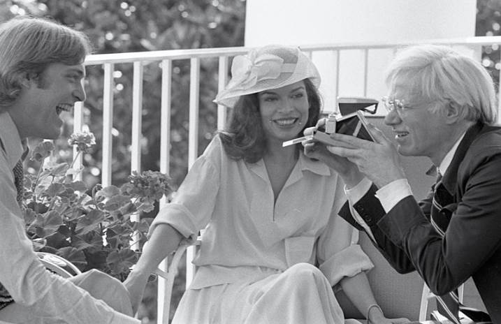 Photograph of Andy Warhol Taking a Polaroid Picture while Sitting with Jack